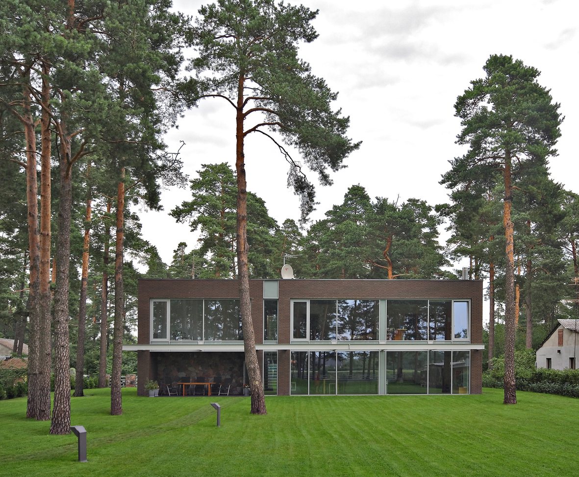 Two-storey brick house with floor-to-ceiling glass, surrounded by tall pine trees on a green lawn